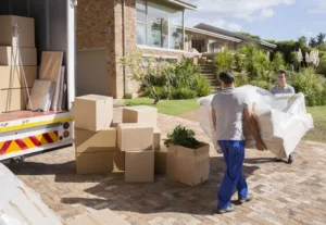Movers carrying packed furniture outside a villa with boxes stacked near a truck